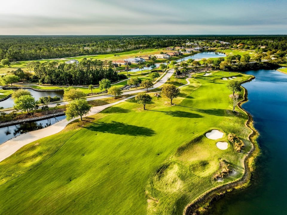Aerial view of a golf course fairway with a water hazard, sand bunkers, and tree-lined residential buildings beyond