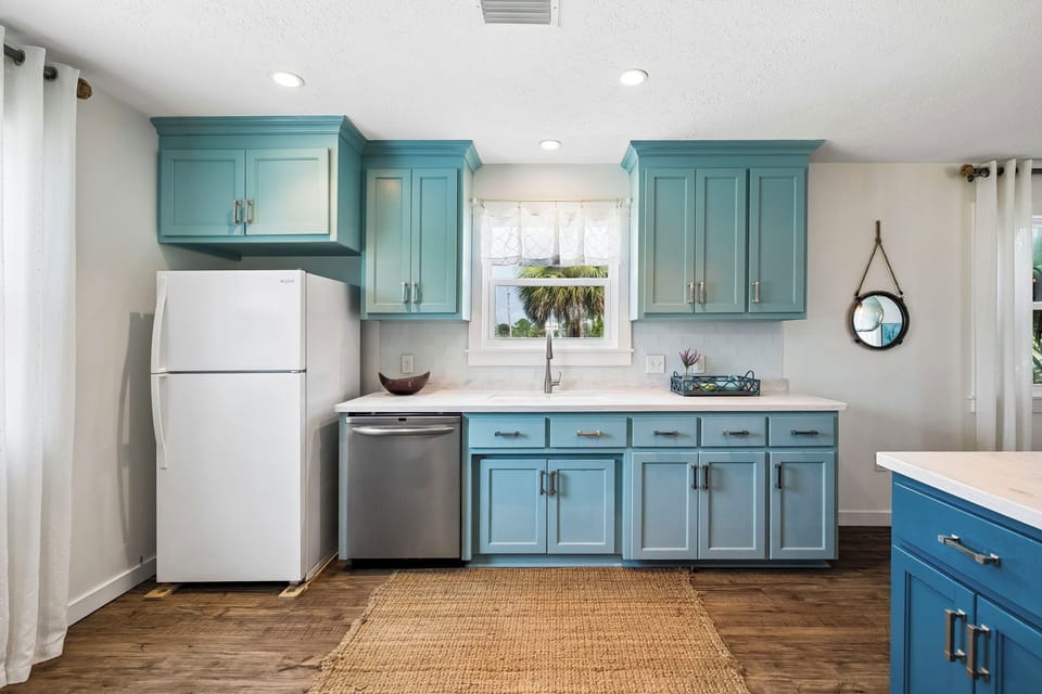 Fully equipped kitchen with coastal-blue cabinetry and modern appliances. Natural light streams through windows overlooking swaying palms, creating a bright and tropical cooking space.