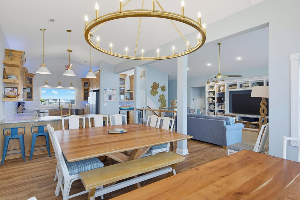 Step into a bright open-concept living space where coastal charm meets comfort. The grand chandelier draws your eye across warm wood floors, connecting your dining area with the cozy lounge—perfect for gathering after a day at the beach.