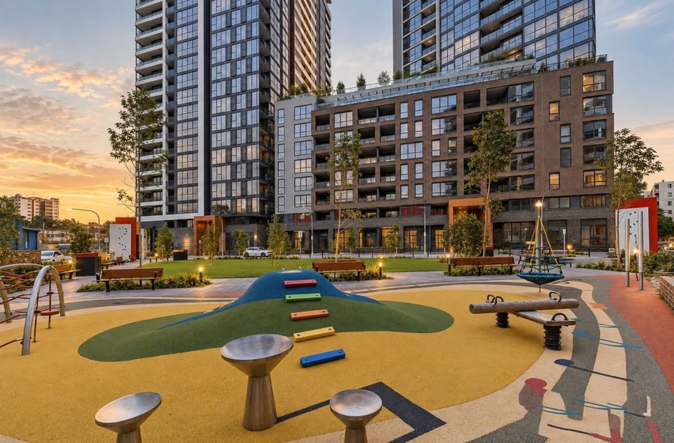 Shared building playground at sunset with a climbing mound, spring play equipment, a nest swing and outdoor percussion instruments on colourful rubber flooring. Residential towers behind. This is a shared building amenity.