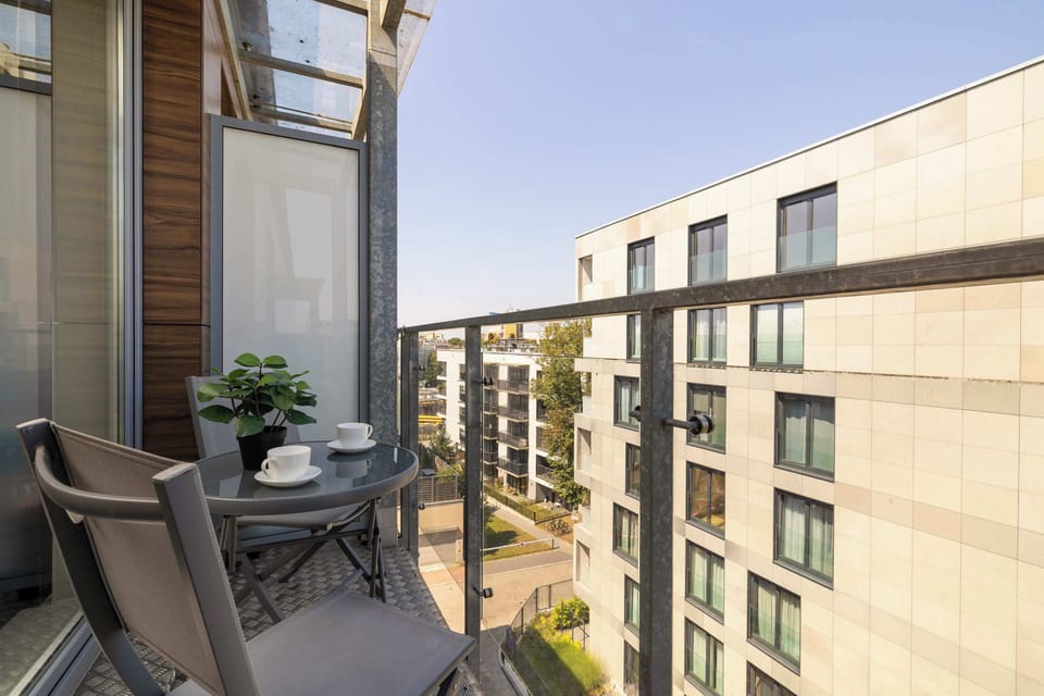 A balcony setting featuring a small table and two dark chairs ready for use. The background reveals a modern residential building complex basking in sunlight.