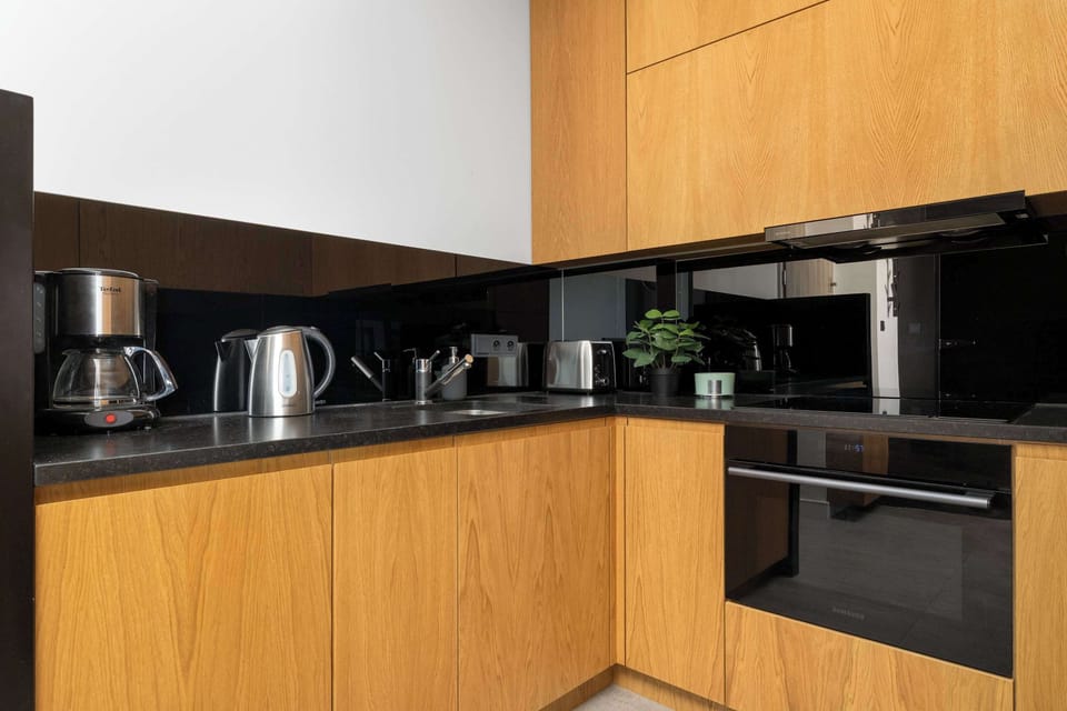 A modern kitchenette area with light wooden cabinetry and a glossy black backsplash. A coffee maker, kettle, and built-in oven sit neatly on the countertops.