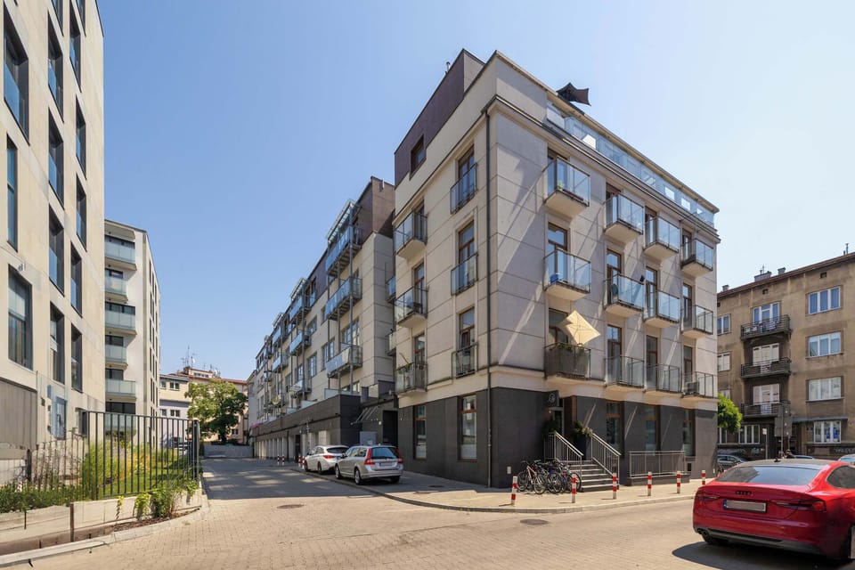 An exterior view of a multi-story apartment building with multiple balconies and a dark gray ground floor. Cars are parked neatly along the street on a bright, sunny day.