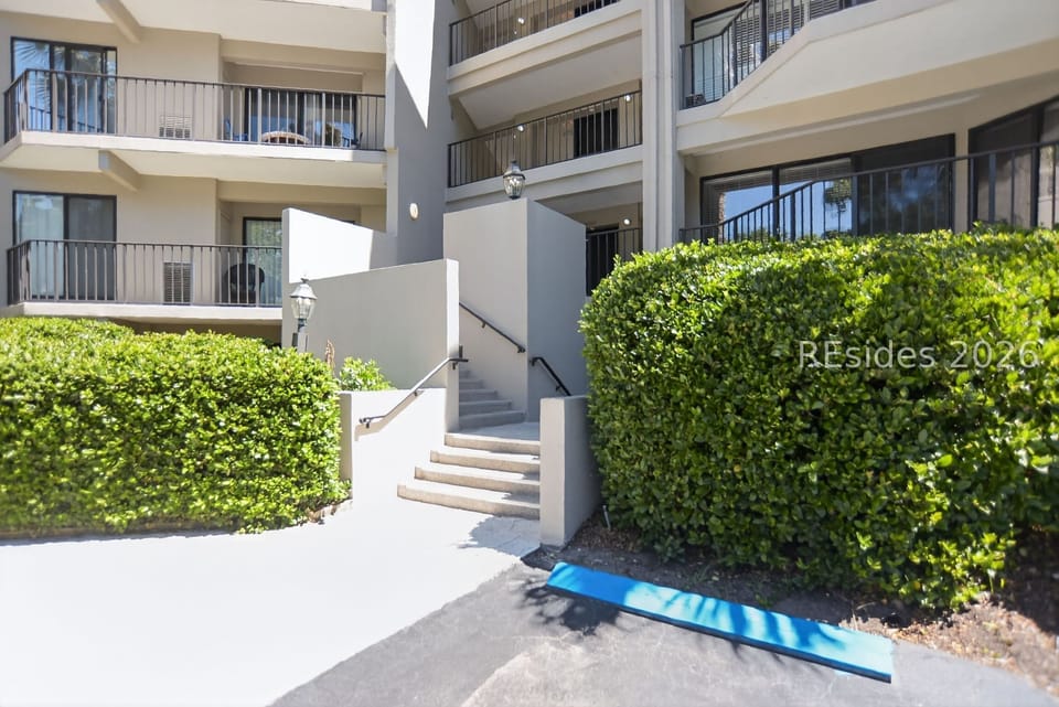 Lush greenery frames the clean architectural lines of this sunlit entryway, where stone steps lead toward private balconies. Bright midday rays catch the minimalist stucco and black railings, creating a calm and orderly path home.