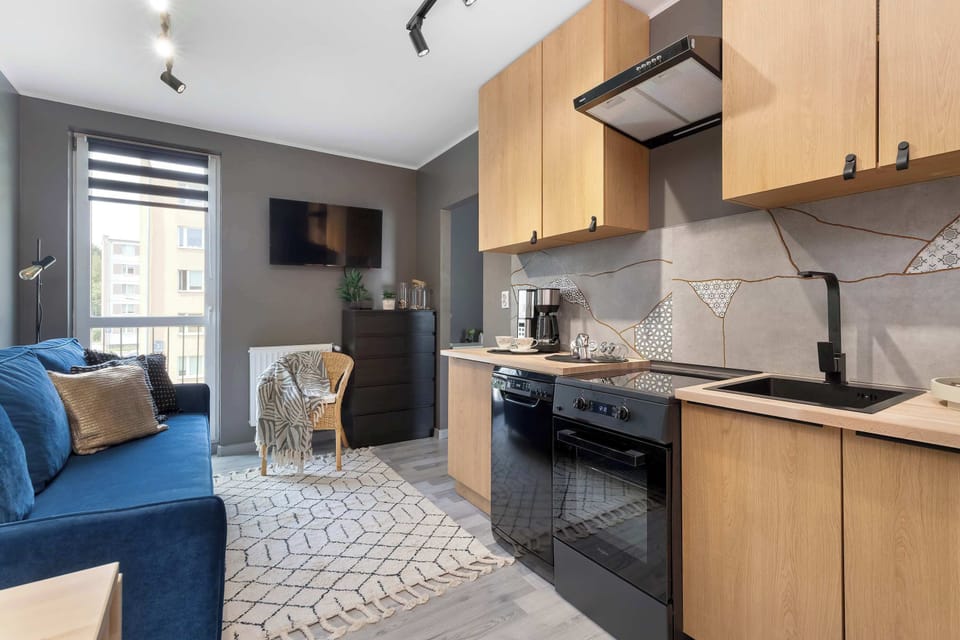 A broad view reveals a fully equipped kitchenette merging into a bright living area. The warm wooden cabinets contrast nicely with the bold blue sofa and dark modern appliances.