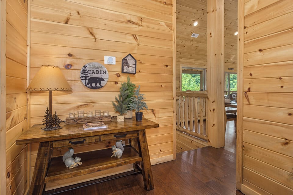 Step inside your cozy cabin retreat where warm wood planks embrace every surface. The rustic console welcomes you with charming forest-inspired décor, while natural light streams through windows framing lush greenery beyond.