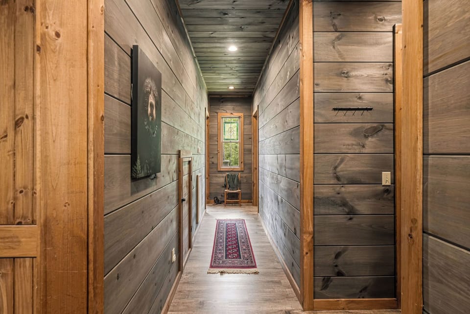 A warm hallway with rustic wood-paneled walls and ceiling leads to a peaceful nook, complete with a cozy reading chair and colorful runner beneath your feet.