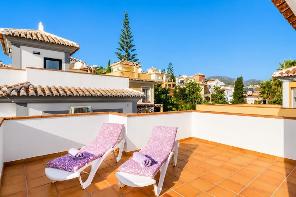 Private rooftop terrace at a modern vacation villa, showcasing a crystal-clear plunge pool, comfortable sun loungers with patterned purple cushions, a wooden pergola, and scenic views of a Mediterranean-style resort town.