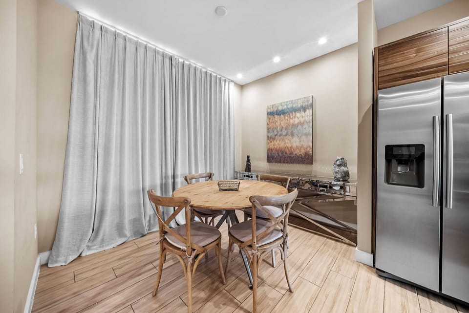 This bright and welcoming dining area boasts a round wooden table with stylish chairs, ideal for shared meals. Soft natural light through elegant curtains adds warmth, creating a cozy atmosphere for all to enjoy.