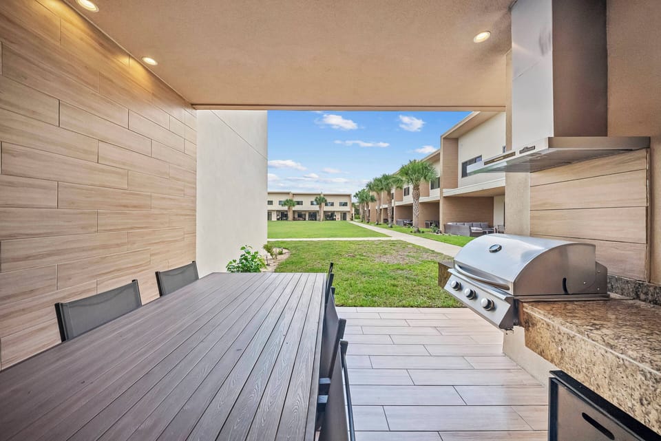 Enjoy our charming outdoor dining area with a sleek wooden table perfect for al fresco meals. The built-in grill and granite countertop create an ideal setting for summer barbecues, surrounded by lush greenery and blue skies.