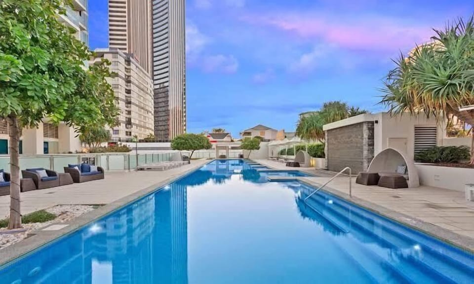Take a refreshing dip in this long, sparkling outdoor lap pool, lined with comfortable sun loungers, cozy poolside cabanas, and tropical palm trees, all set against a backdrop of modern high-rise buildings.