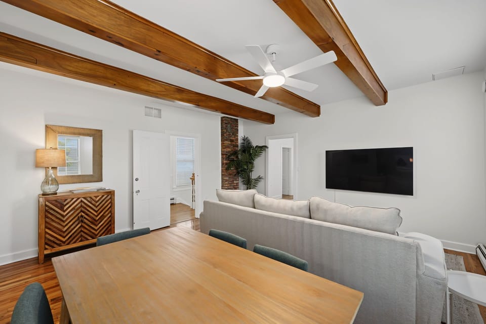 This cozy view of the second-level living space showcases warm exposed wood ceiling beams, a large wall-mounted flat-screen TV framed by a plush sofa and an adjacent dining table.