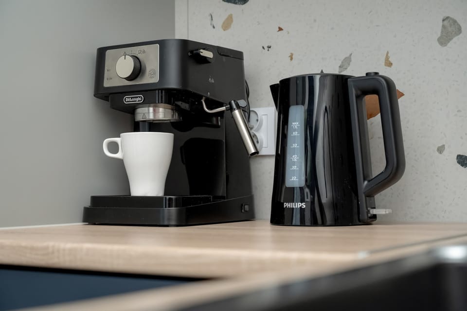 A close-up detail of small kitchen appliances placed on a light wooden countertop. An espresso machine and a black electric kettle stand ready for morning coffee or tea.