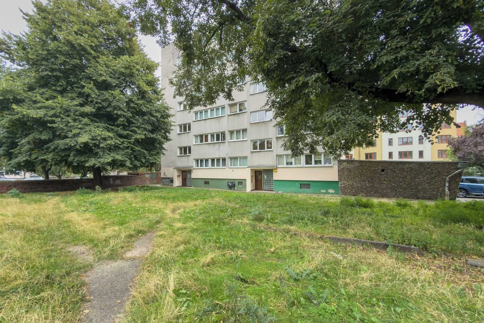 An exterior view of a multi-story apartment building surrounded by green grass and mature trees. The peaceful residential grounds offer a pleasant green setting.