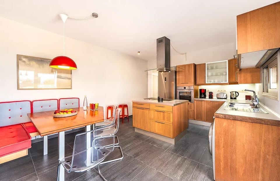 A modern kitchen features wooden cabinetry, a central island with an overhead range hood, and an eat-in dining area with a red cushioned bench and clear chairs.