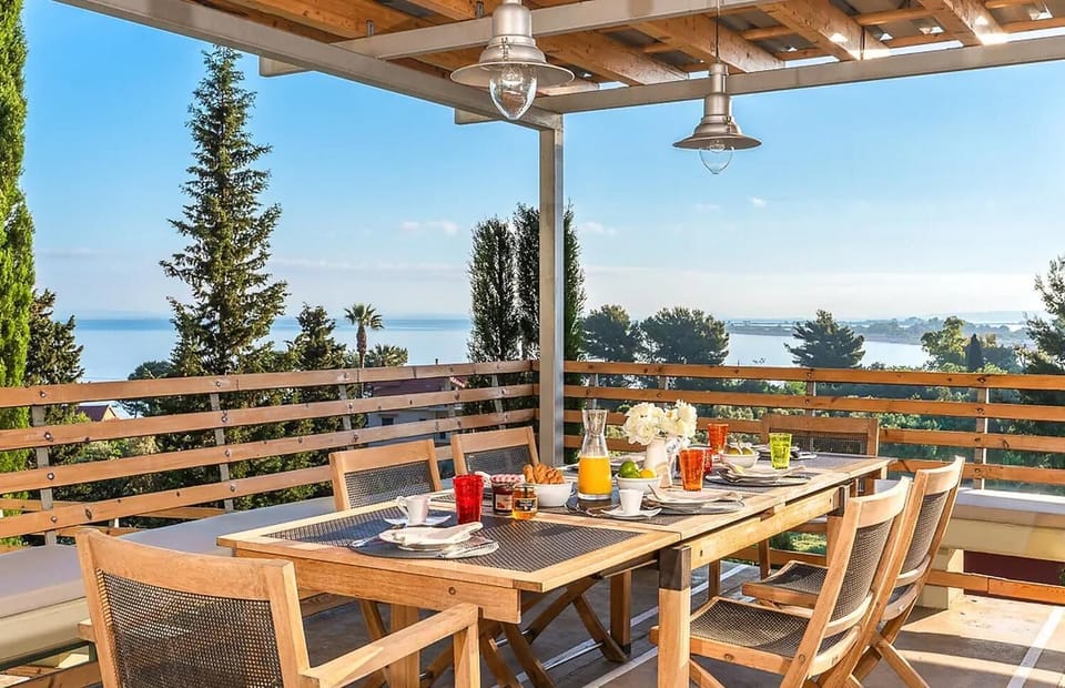 A wooden outdoor dining table is set for breakfast on a covered terrace, offering a panoramic view of the sea and surrounding trees.