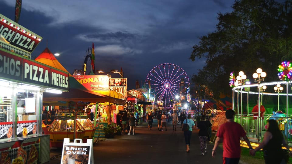 Nighttime fair scene with food stands and a Ferris wheel.