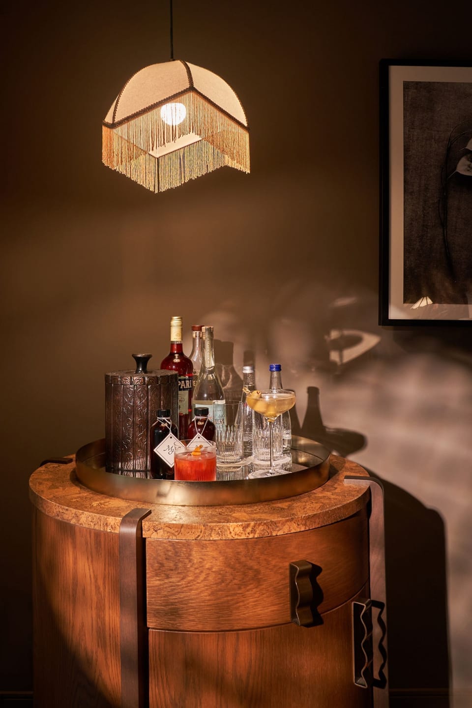 Bar cart with a round metal tray, assorted glass bottles, and a gold fringe lampshade, set against framed artwork