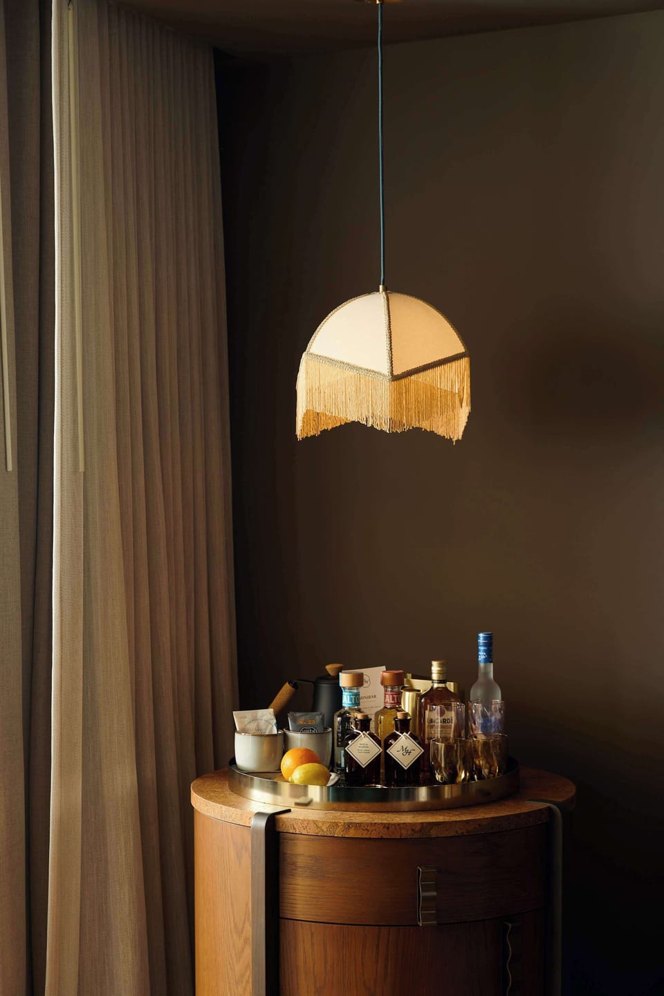 Round wooden bar cart with liquor bottles and a lemon beneath a fringed pendant lamp, against a dark brown wall