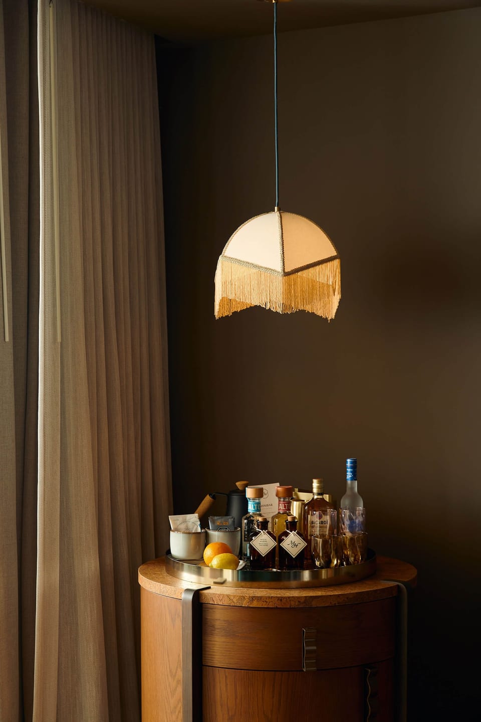 Walnut bar cabinet topped with spirits, bitters, and glassware under a gold fringe pendant lamp