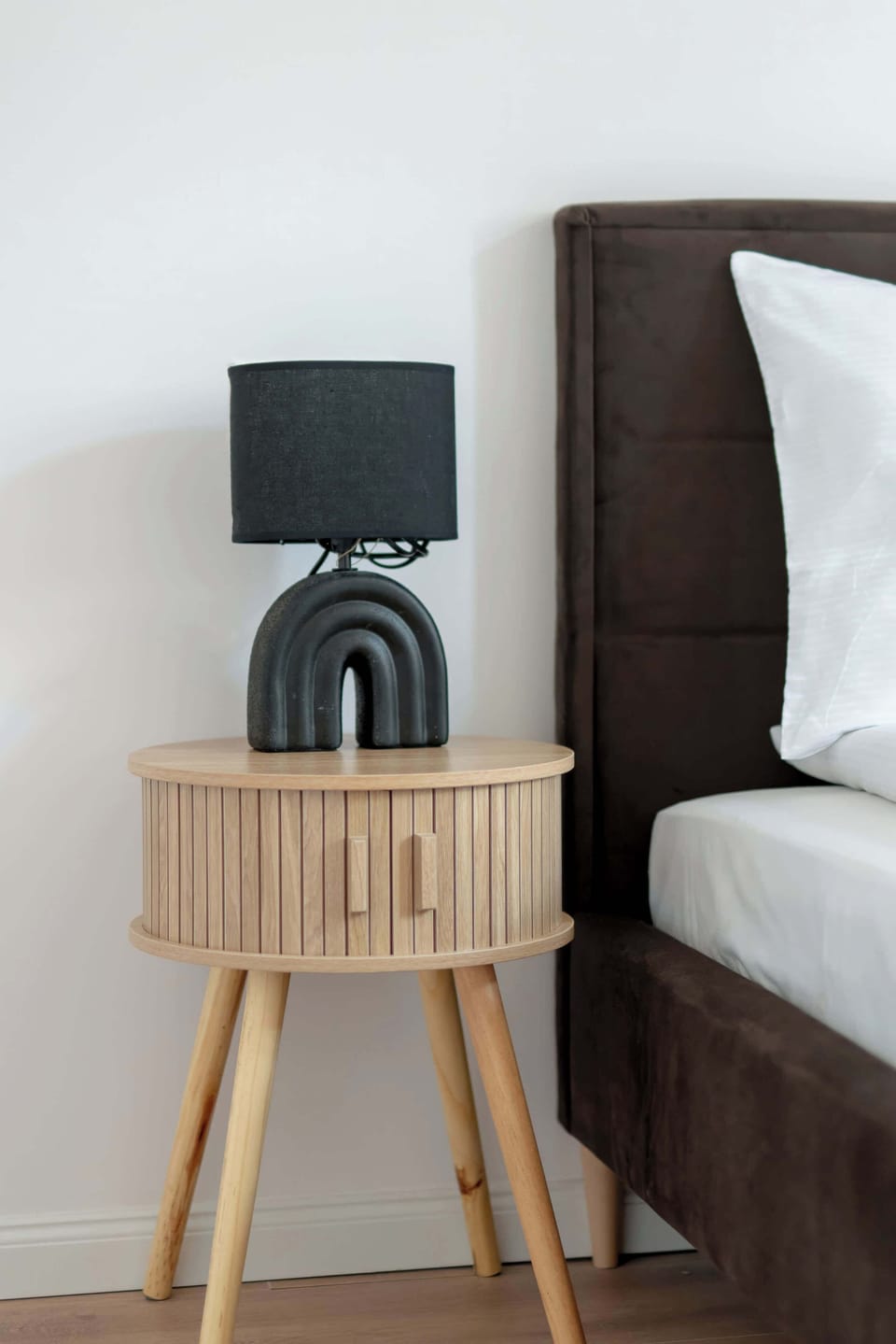 A detailed shot focuses on a round wooden bedside nightstand holding a black table lamp next to a dark upholstered bed frame.