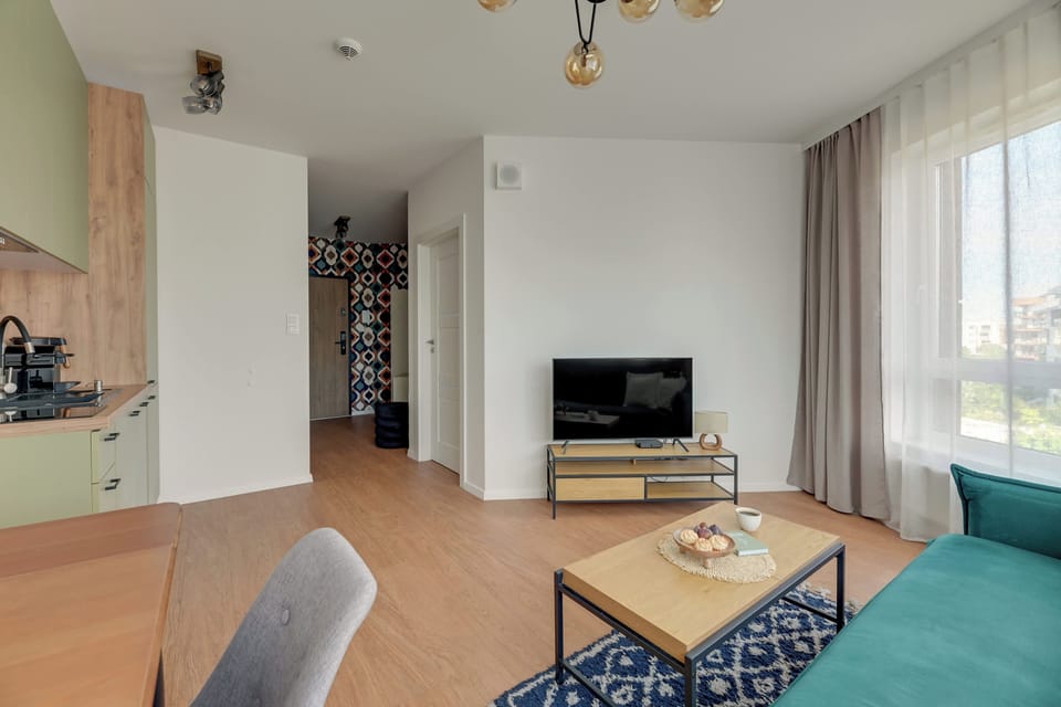 Jasny i przytulny salon z wygodną sofą, elegancką strefą wypoczynku i dużymi przeszkleniami zapewniającymi naturalne światło. Turkusowe akcenty, dekoracyjny dywan i harmonijna aranżacja nadają wnętrzu świeży, nowoczesny charakter.