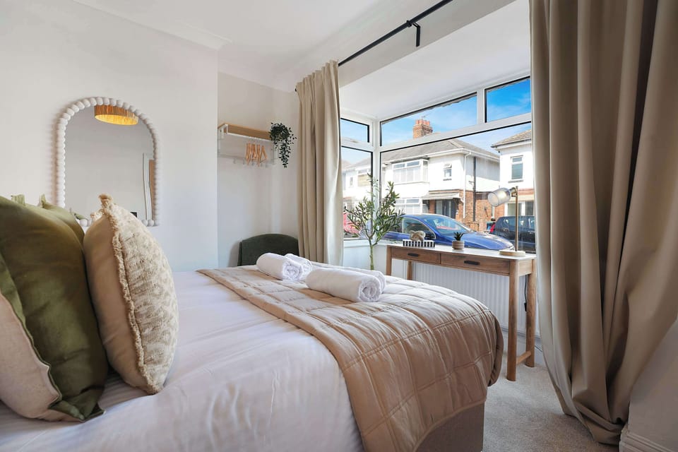 This spacious double bedroom features a comfortable double bed with two bedside tables and matching lamps. Soft furnishings create a welcoming atmosphere, while a wall-mounted mirror, a chair, a hanging rail, and a table by the window add comfort. 