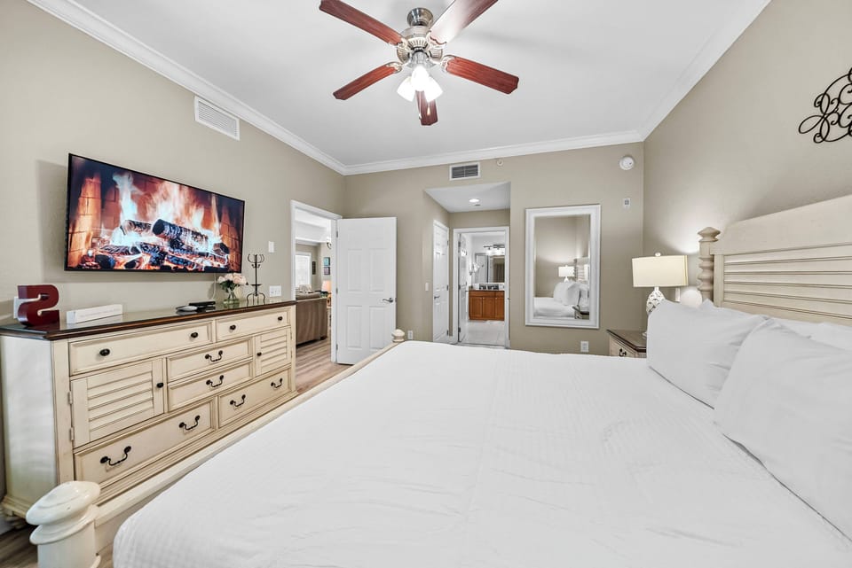 Welcome to a cozy bedroom with a spacious king bed dressed in crisp white linens, a charming wooden dresser, and a stylish ceiling fan. Warm neutral tones create a welcoming vibe, and the large TV offers a touch of modern comfort.
