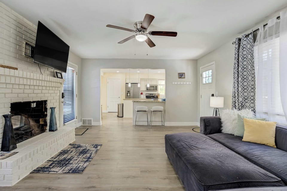 A wide opening connects this living area directly to the kitchen, with bar seating visible just beyond. The white painted brick fireplace extends to a raised hearth, and a patterned rug grounds the seating over light plank floors.