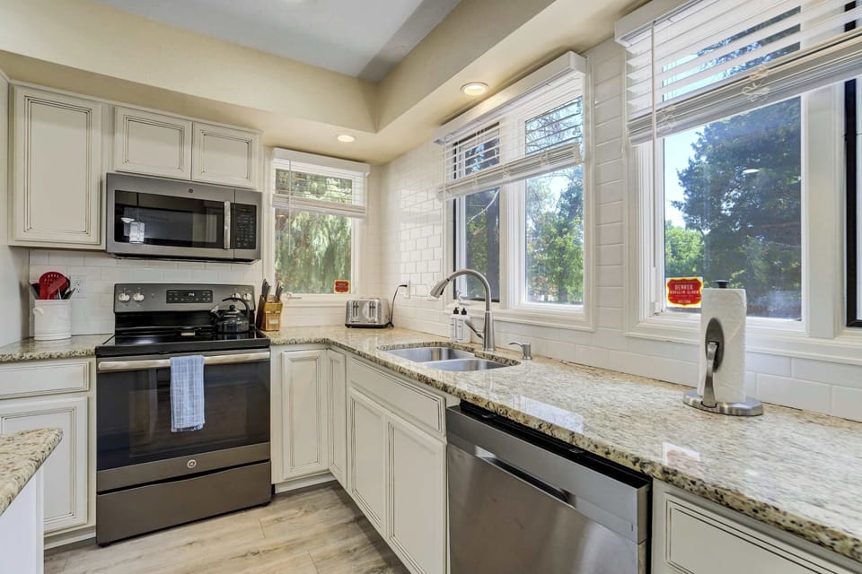 Stainless appliances line the cooking wall, with a full range and over range microwave set into cream cabinetry. A bank of windows floods the counters with daylight, making the whole space bright and easy to cook in together.