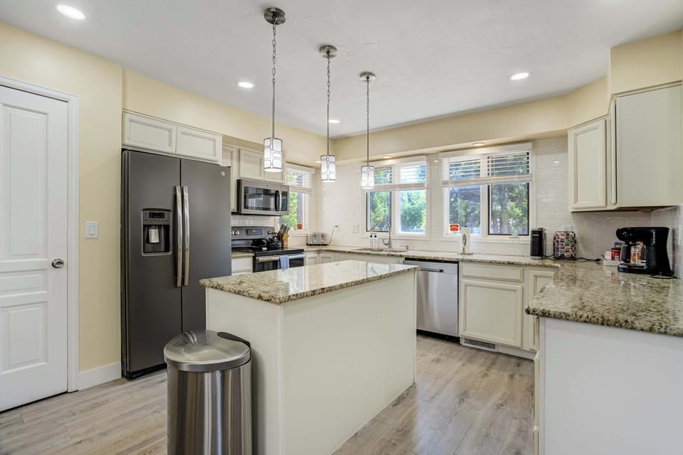 Speckled granite counters wrap a center island with room to prep and gather, framed by cream cabinetry and stainless appliances throughout. Windows above the sink bring steady natural light into the heart of the kitchen.