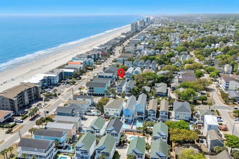 An aerial view captures a charming beachfront neighborhood where coastal homes nestle among lush greenery, just steps from the sandy shore and sparkling ocean.