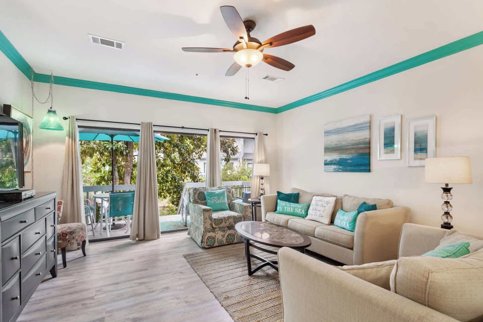 Coastal-chic living room with teal crown molding, beachy throw pillows, and sliding doors opening to a shaded patio. 35 Ocean Breeze brings breezy Hilton Head style home.