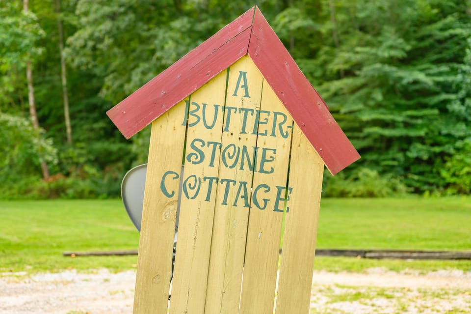 Wooden sign shaped like a house with red roof and blue lettering reading "A Butterstone Cottage" against green trees.