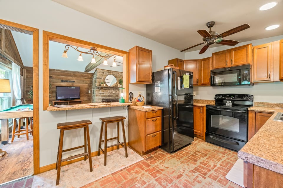 Kitchen features black appliances, warm wood cabinets, brick-patterned tile floor, and a pass-through with two wooden stools.