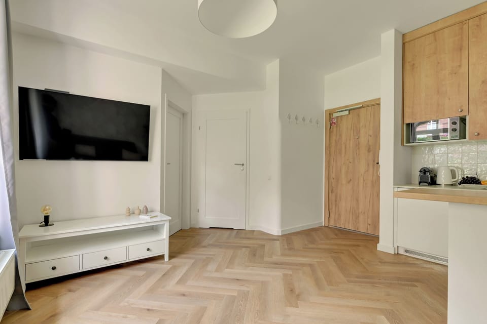 Jasny i przytulny salon z wygodną sofą, dużymi oknami i elegancką podłogą ułożoną w jodełkę. Delikatne dekoracje i stonowana kolorystyka tworzą harmonijną, komfortową przestrzeń do wypoczynku.
