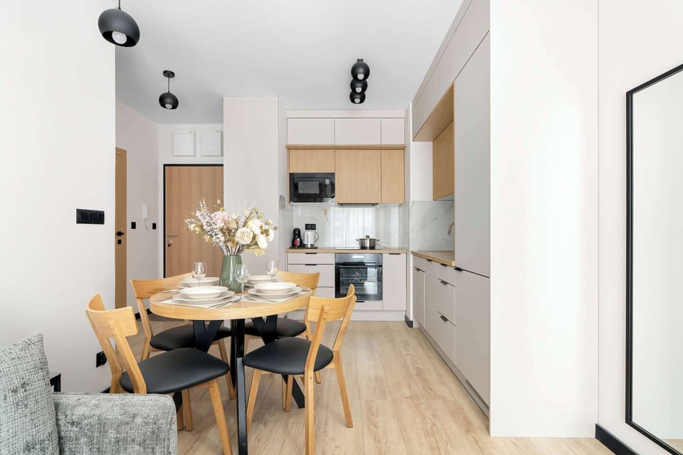 A stylish open-plan dining space featuring a round table surrounded by wooden chairs. A decorative vase of pink flowers serves as a tabletop centerpiece, leading into the adjacent kitchen area.