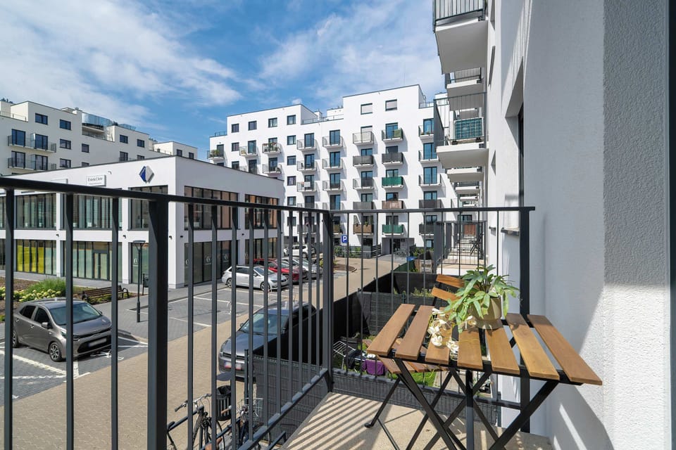 A outdoor balcony set with a small wooden bistro table and chairs, enclosed by black metal railings. Surrounding modern apartment buildings frame the sunny exterior view.