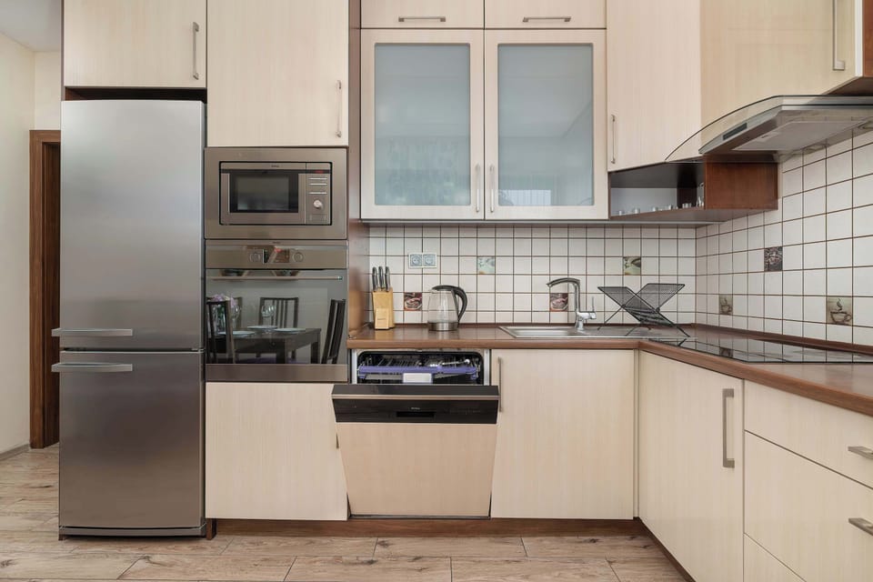 A modern kitchen with a refrigerator, oven, microwave, and light wooden cabinetry. The large window provides natural light and makes the space feel open.