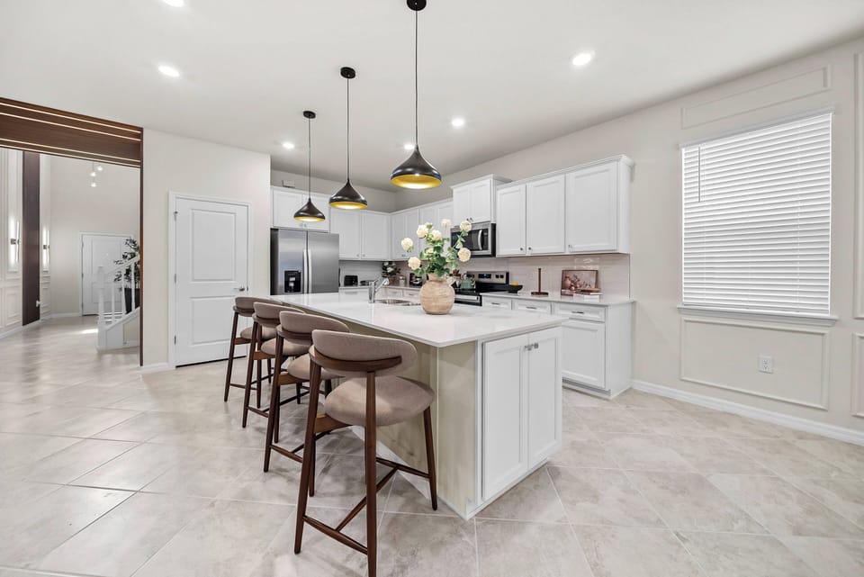Experience a bright, spacious kitchen with sleek white cabinetry and a large central island, ideal for casual dining or meal prep. Elegant pendant lights and a big window enhance the warm ambiance and inviting atmosphere.