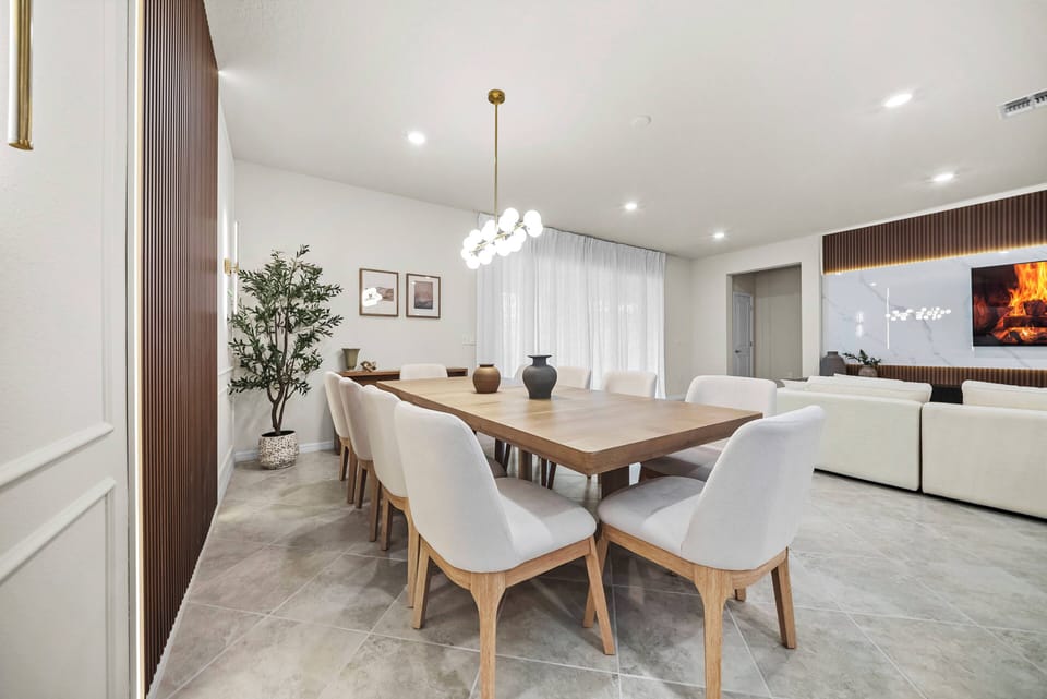 Join us for communal dining at a spacious wooden table, surrounded by sleek white chairs and warm decor. The soft glow of pendant lights creates an inviting atmosphere, complemented by an elegant plant for a natural touch.