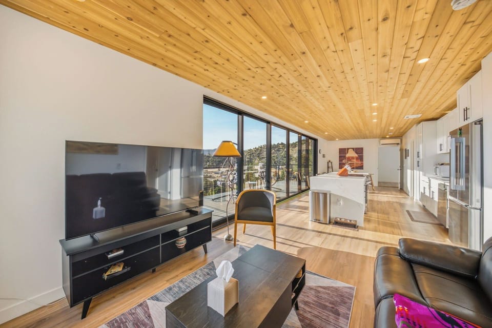 An open concept living room and kitchen with a large TV, comfortable seating, and a view of the outdoors through expansive windows.