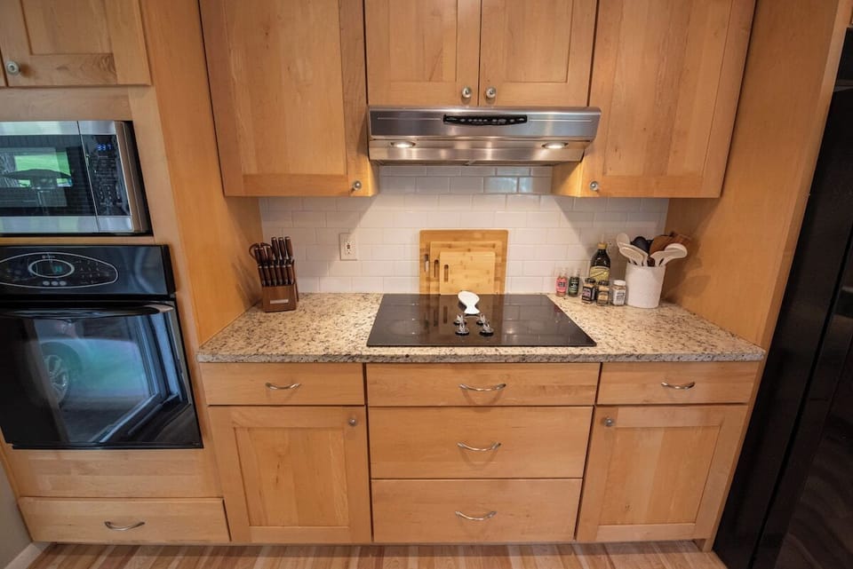 Prepare meals efficiently in this kitchen cooking zone featuring a black glass cooktop with an overhead range hood, natural wood cabinetry, built-in wall oven and microwave, granite-style countertops, and a white subway tile backsplash.
