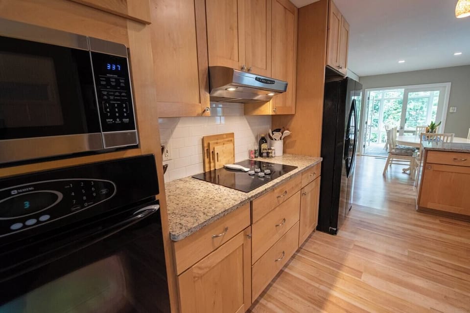 Prepare meals with ease in this well-equipped kitchen featuring built-in appliances, a glass stovetop with a vent hood, natural wood cabinetry, granite-style countertops, and light wood flooring leading toward an open dining area.