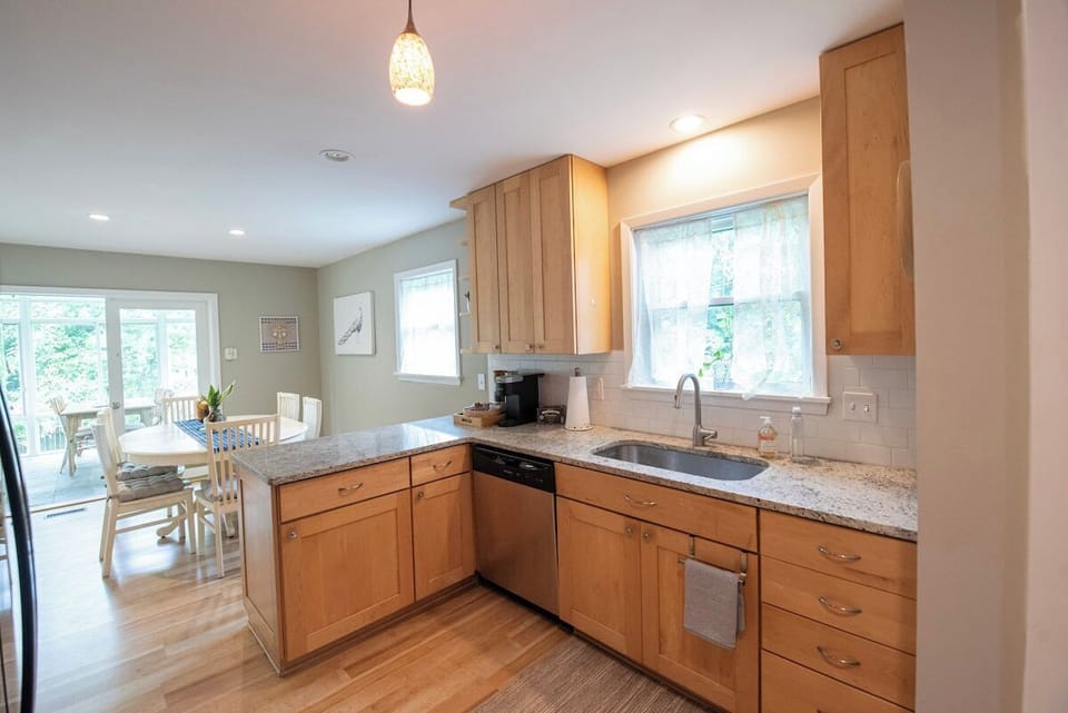 Prepare meals in this open kitchen featuring light wood cabinetry, granite-style countertops, an undermount sink under a sunny window, a dishwasher, and a seamless view into the adjacent dining area.