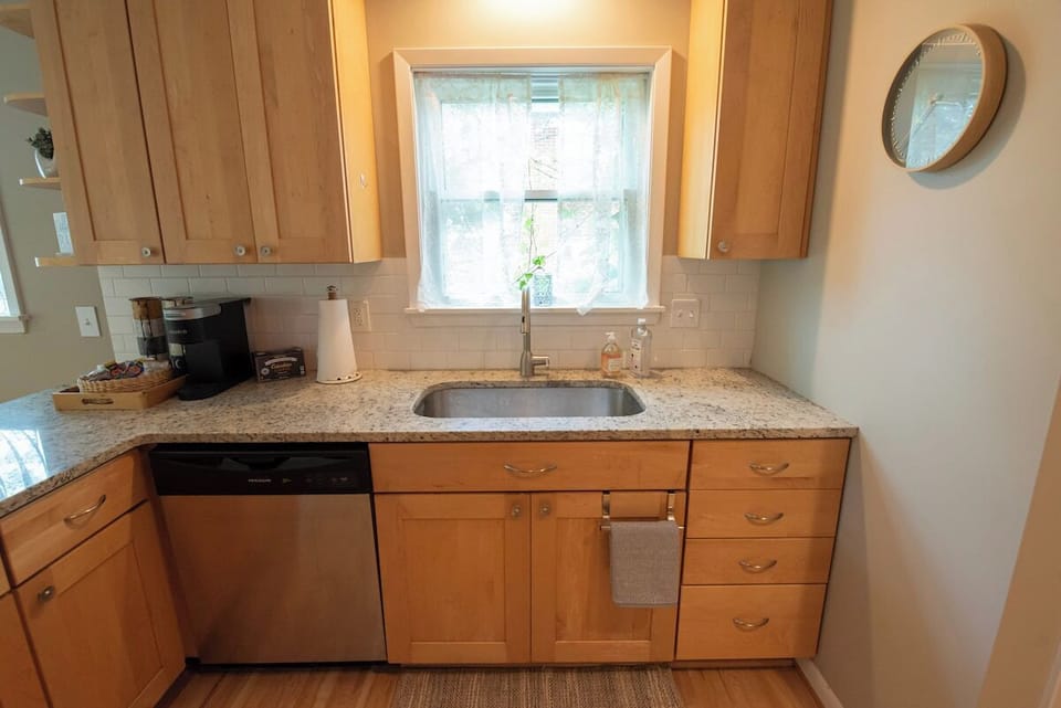 Prepare meals with ease in this bright kitchen area featuring light wood cabinetry, granite-style countertops, an undermount stainless steel sink positioned beneath a window, a dishwasher, and a small round wall mirror.