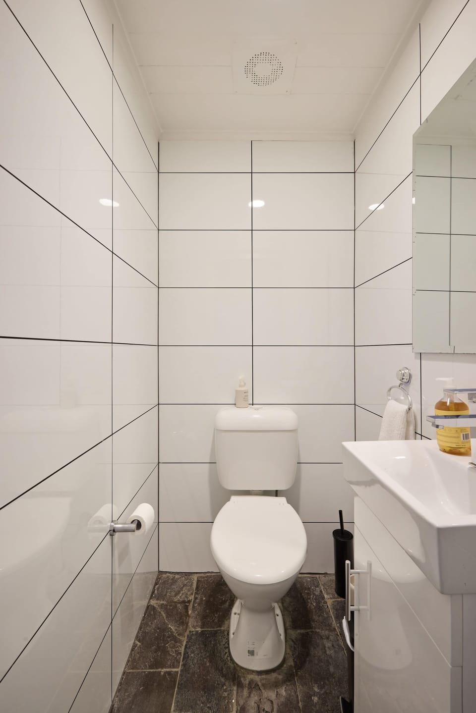Observe this compact powder room fitted with full-height white wall tiles, dark floor tiles, a dual-flush toilet, and a small side-mounted vanity.