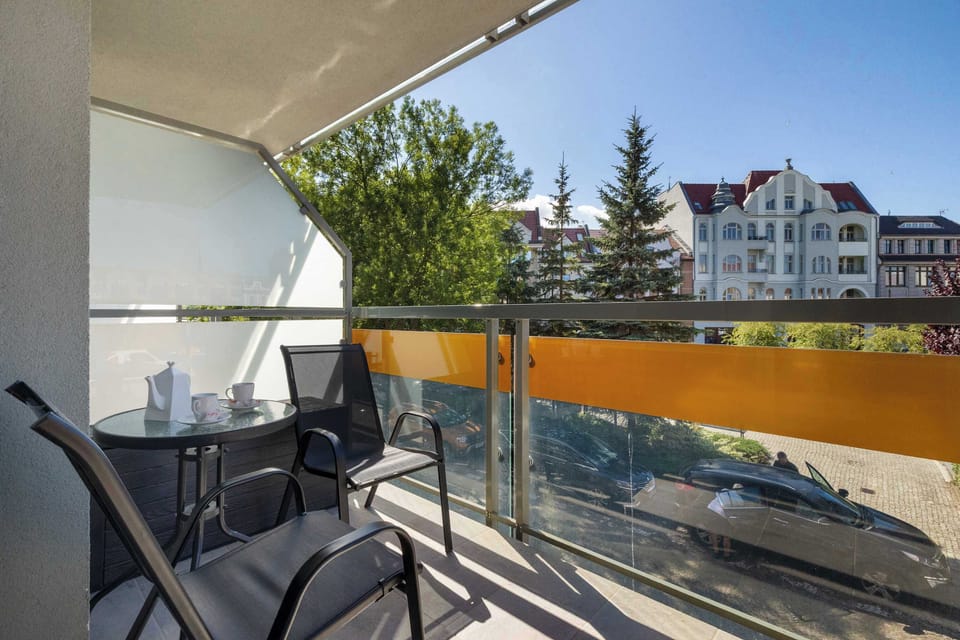Outdoor balcony space looking out over green tree canopy and local architecture. A patio table and two chairs are set up near the metal railings.