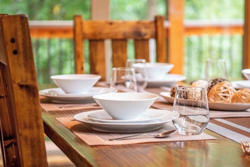 Your dining table is beautifully set with crisp white dishes and rustic wooden chairs, framed by large windows overlooking lush greenery—a perfect spot to enjoy morning coffee or a leisurely meal together.