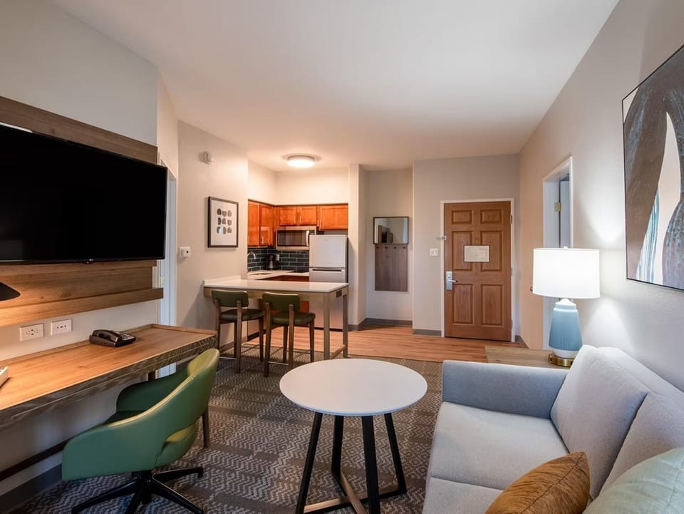 Living area with gray sofa, work desk, flat-screen TV, and kitchen with dining bar in view